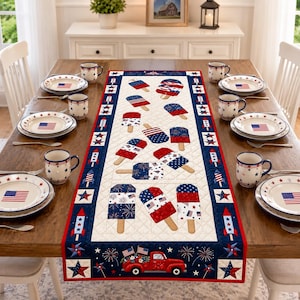 May include: A rectangular quilted table runner featuring a patriotic theme with red, white, and blue ice lolly patterns, rocket motifs, and a red truck carrying American flags. The runner is centred on a wooden dining table with matching plates.