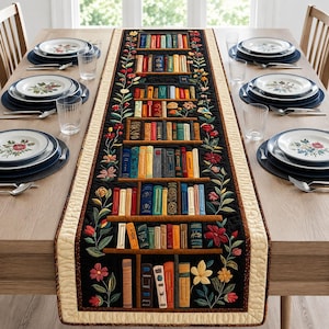 Puede incluir: Camino de mesa negro con diseño de estantería con libros coloridos y motivos florales. El camino de mesa tiene un borde color crema y está colocado sobre una mesa de madera.