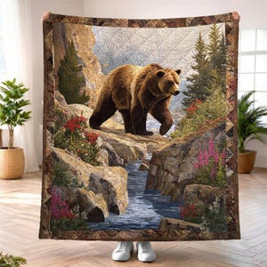 Puede incluir: Un edredón marrón y beige con una imagen detallada de un oso pardo caminando por la orilla rocosa de un río. El edredón tiene un diseño de paisaje con árboles, flores y una cascada. El borde es marrón oscuro con un patrón geométrico.