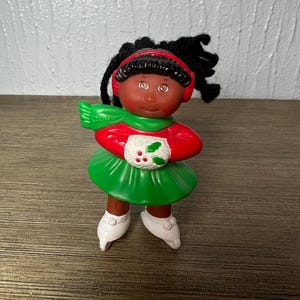 May include: A Cabbage Patch Kids figurine featuring a doll with dark skin, brown eyes, and black yarn hair. The doll wears a red long-sleeved top, a green skirt, a green scarf with CPK text, red earmuffs, and white ice skates while holding a white muff.