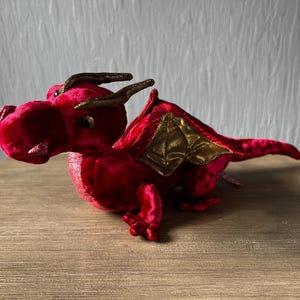 May include: A plush red dragon toy with gold-colored accents on the wings and horns. The dragon has a textured, velvet-like appearance and is posed on a wooden surface. The dragon's eyes are small and dark.