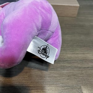 Puede incluir: Un peluche de color lavanda con una etiqueta de Disney adjunta. La etiqueta es blanca con el logotipo de Disney en negro. El peluche es suave y parece estar hecho de un material aterciopelado. El fondo es una superficie de madera.