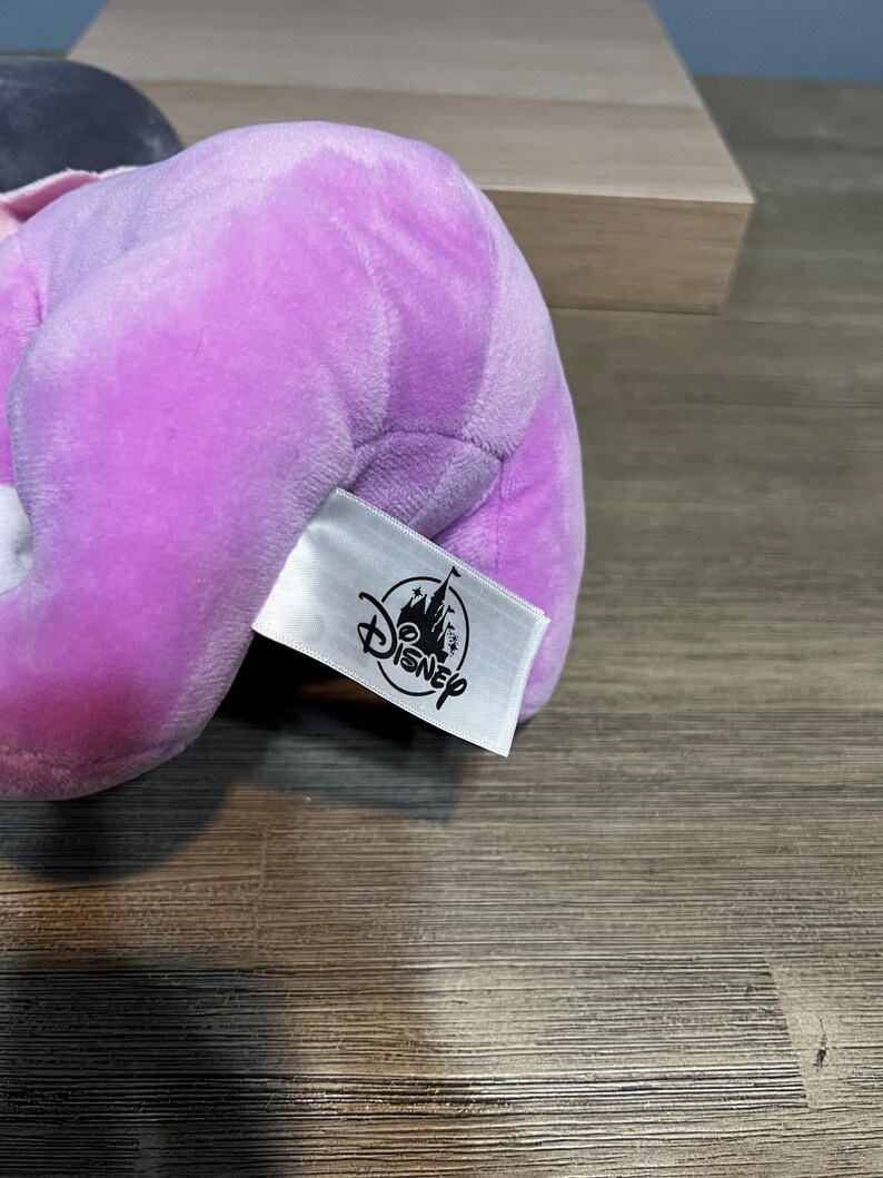 Puede incluir: Un peluche de color lavanda con una etiqueta de Disney adjunta. La etiqueta es blanca con el logotipo de Disney en negro. El peluche es suave y parece estar hecho de un material aterciopelado. El fondo es una superficie de madera.