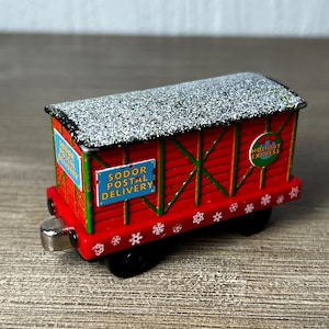 May include: A red toy train cargo car with green trim and a silver roof featuring a dusting of white snow. The side displays blue rectangular signs reading SODOR POSTAL DELIVERY and a circular green logo reading HOLIDAY EXPRESS with white snowflakes.