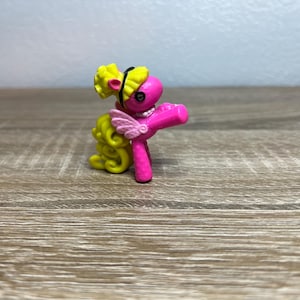 May include: A small pink pony figurine with a bright yellow curly mane and tail. The pony features a black headband, a white pearl-like necklace, and small light pink wings on its back. The figure is standing in a pose with one front leg raised.