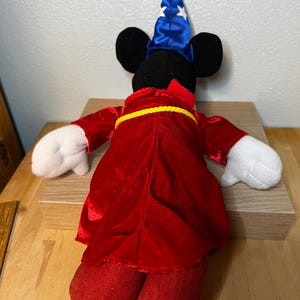 K&ouml;nnte beinhalten: Ein Pl&uuml;sch-Mickey Mouse als Zauberer verkleidet, mit einem blauen Hut mit wei&szlig;en Sternen und einer roten Robe mit gelbem G&uuml;rtel. Das Spielzeug hat schwarze Ohren, wei&szlig;e Handschuhe und eine rote Hose. Das Spielzeug steht auf einer Holzoberfl&auml;che.