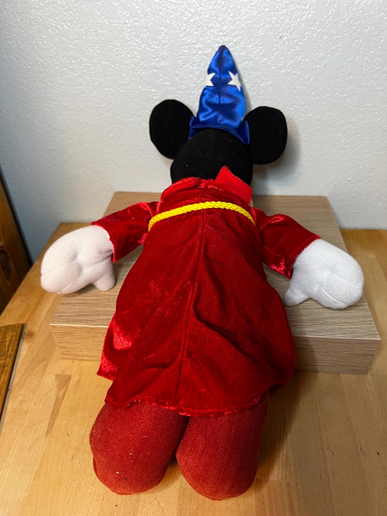K&ouml;nnte beinhalten: Ein Pl&uuml;sch-Mickey Mouse als Zauberer verkleidet, mit einem blauen Hut mit wei&szlig;en Sternen und einer roten Robe mit gelbem G&uuml;rtel. Das Spielzeug hat schwarze Ohren, wei&szlig;e Handschuhe und eine rote Hose. Das Spielzeug steht auf einer Holzoberfl&auml;che.