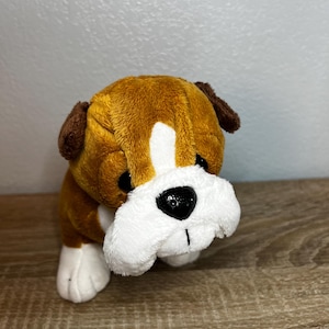 May include: A plush bulldog toy featuring soft brown and white fur, dark brown ears, and a prominent white patch extending from the forehead to the snout. The stuffed animal has large black plastic eyes and a shiny black nose, with white paws.