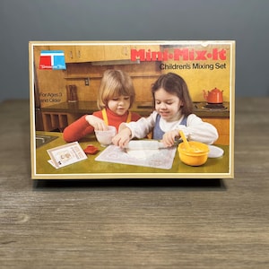 May include: A vintage Tupperware Mini-Mix-It Children's Mixing Set box. The box features an illustration of two children using the set. The box has the Tupperware logo and text that reads "For Ages 3 and Older". The box is on a wooden surface.