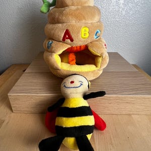 May include: A plush toy set featuring a tan beehive with the letters A, B, and C, and a smiling bee with yellow and black stripes, red wings, and black legs. The beehive has a green stem and an opening in the front.