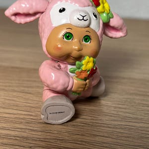 May include: A pink figurine of a lamb-like character with a human-like face, green eyes, and a flower crown. The figurine is holding a small bouquet of flowers. The base of the figurine has the text "CPW0860 061322-HK".