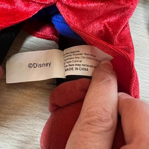 K&ouml;nnte beinhalten: Ein Pl&uuml;schtier mit rotem, gelbem und blauem Stoff. Auf dem Etikett steht "&copy;Disney" und "MADE IN CHINA". Das Spielzeug besteht aus Polyesterfasern und ist f&uuml;r Kinder ab 3 Jahren geeignet.