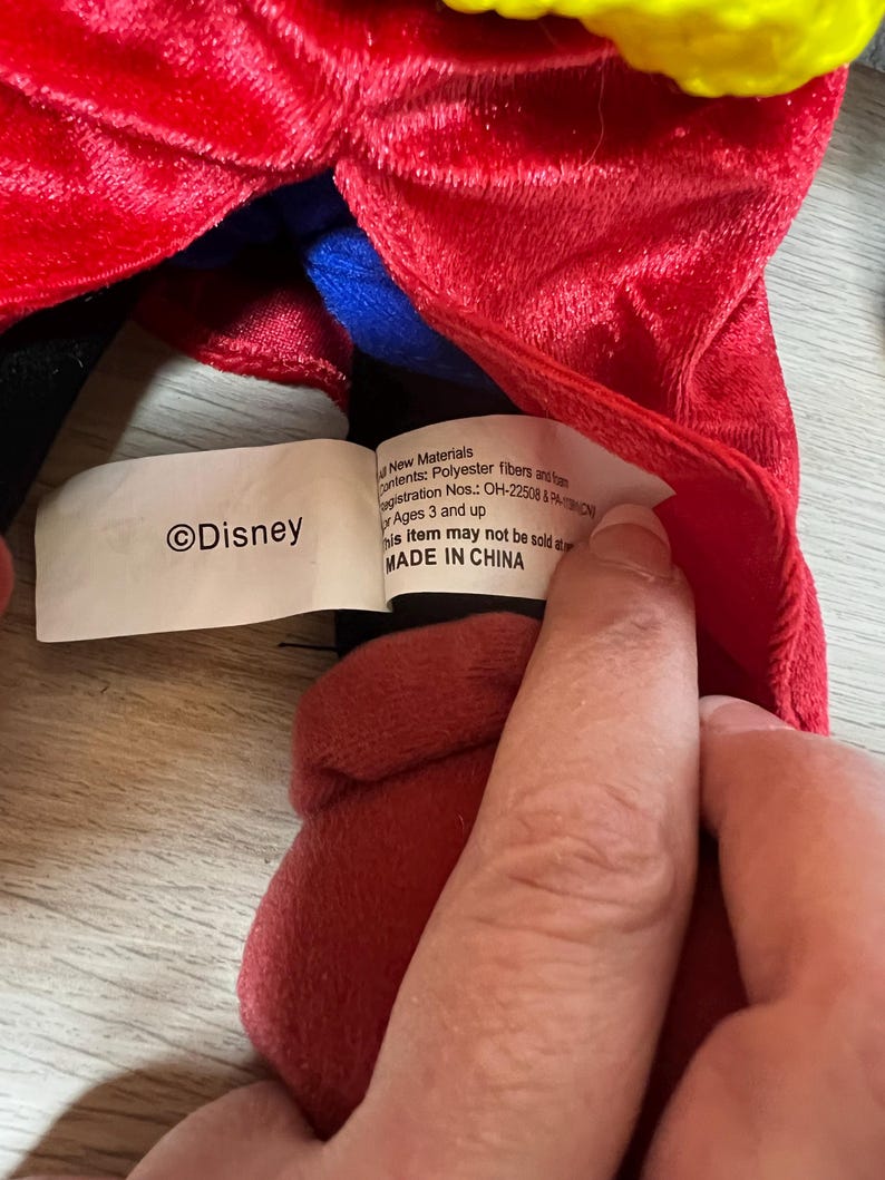 K&ouml;nnte beinhalten: Ein Pl&uuml;schtier mit rotem, gelbem und blauem Stoff. Auf dem Etikett steht "&copy;Disney" und "MADE IN CHINA". Das Spielzeug besteht aus Polyesterfasern und ist f&uuml;r Kinder ab 3 Jahren geeignet.