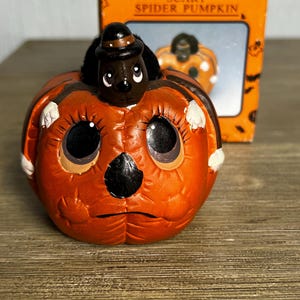 May include: An orange pumpkin figurine with large black eyes and a black nose, featuring a small black spider wearing a hat on top. The pumpkin is positioned in front of its original orange cardboard box, which displays the text SCARY SPIDER PUMPKIN.