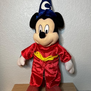 K&ouml;nnte beinhalten: Ein Pl&uuml;sch-Mickey Mouse als Zauberlehrling aus Fantasia. Die Figur tr&auml;gt eine rote Robe mit gelbem Seilg&uuml;rtel, rote Schuhe und einen blauen Zauberhut mit wei&szlig;en Sternen und einem Halbmond. Das Spielzeug hat schwarze Ohren und ein cremefarbenes Gesicht.