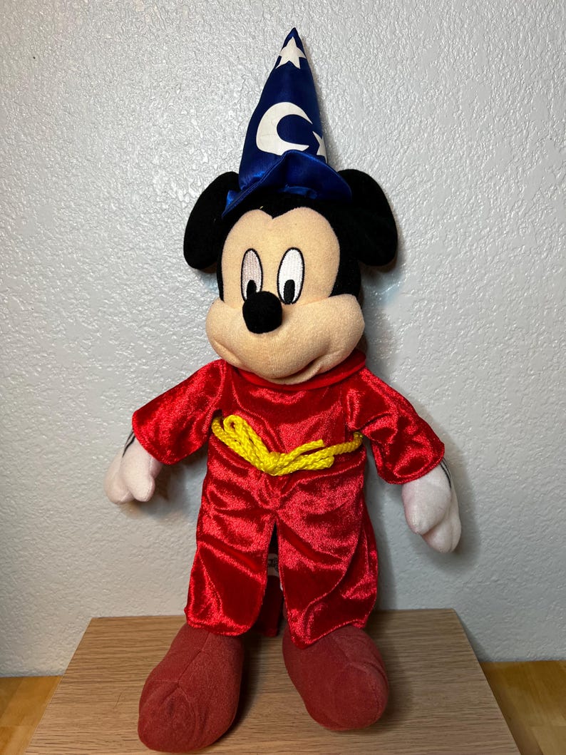 K&ouml;nnte beinhalten: Ein Pl&uuml;sch-Mickey Mouse als Zauberlehrling aus Fantasia. Die Figur tr&auml;gt eine rote Robe mit gelbem Seilg&uuml;rtel, rote Schuhe und einen blauen Zauberhut mit wei&szlig;en Sternen und einem Halbmond. Das Spielzeug hat schwarze Ohren und ein cremefarbenes Gesicht.