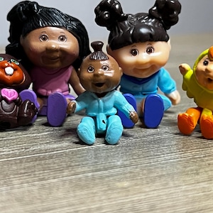 May include: A collection of vintage Cabbage Patch Kids figurines. The set includes dolls with dark hair and various outfits, a brown bear with a heart, and a yellow chick figurine. The dolls are sitting on a wooden surface.