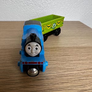 May include: A blue wooden toy train with a smiling face and a silver front. It is connected to a green cargo car with black wheels. The train is on a wooden surface.