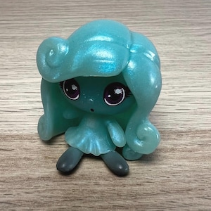 May include: A small, teal-colored figurine with a large head and curly hair. The figurine has large eyes with purple irises and a small, simple dress. The figure is sitting with its legs spread and arms at its sides.