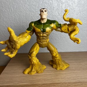 Puede incluir: Una figura de acci&oacute;n de pl&aacute;stico de un personaje musculoso con un torso bronceado, un pecho verde y beige con estampado de camuflaje y brazos y piernas amarillos texturizados. La figura tiene una expresi&oacute;n severa y posa con los brazos extendidos. La figura est&aacute; sobre una superficie de madera.