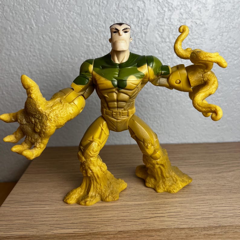 Puede incluir: Una figura de acci&oacute;n de pl&aacute;stico de un personaje musculoso con un torso bronceado, un pecho verde y beige con estampado de camuflaje y brazos y piernas amarillos texturizados. La figura tiene una expresi&oacute;n severa y posa con los brazos extendidos. La figura est&aacute; sobre una superficie de madera.