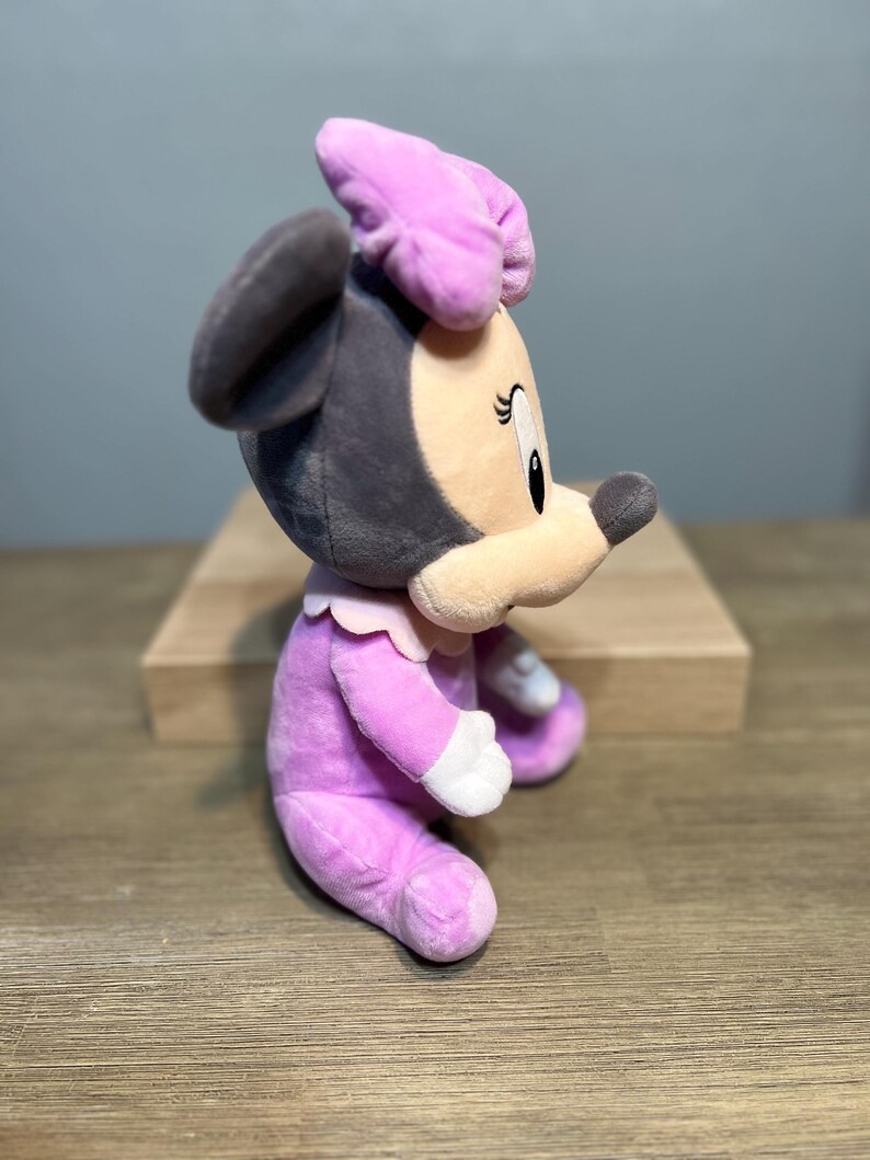 Puede incluir: Mu&ntilde;eca de peluche de Minnie Mouse de perfil. La mu&ntilde;eca lleva un vestido lavanda, un lazo a juego y guantes blancos. La cara es beige con ojos negros y una nariz negra. Las orejas son grises.