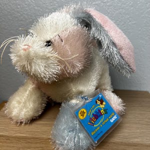 May include: A plush bunny toy with white, pink, and gray fur. The bunny has long ears and a tag that reads "Webkinz" and "Adopt a pet... discover a virtual world!" The toy is designed for children aged 3+.