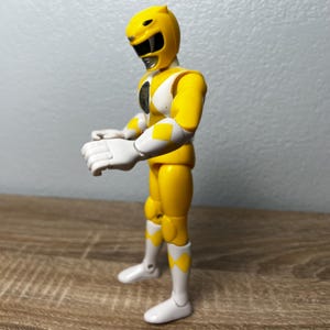 Op de afbeelding: Een gele actiefiguur van een Power Ranger. De figuur draagt een gele helm, handschoenen en laarzen, met witte accenten en ruitvormige gele details. De figuur staat met uitgestrekte armen.