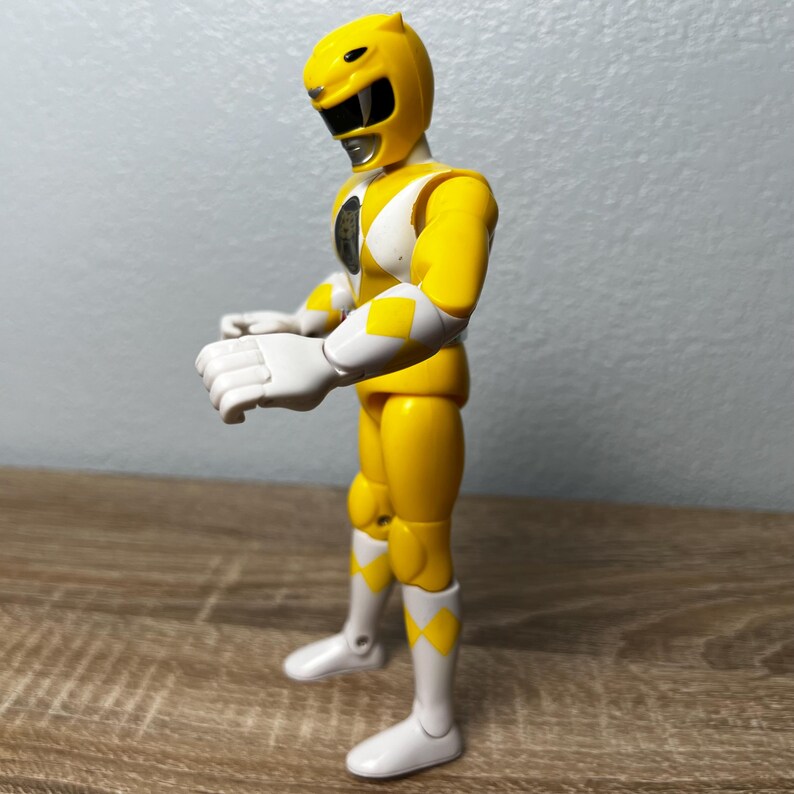 Op de afbeelding: Een gele actiefiguur van een Power Ranger. De figuur draagt een gele helm, handschoenen en laarzen, met witte accenten en ruitvormige gele details. De figuur staat met uitgestrekte armen.