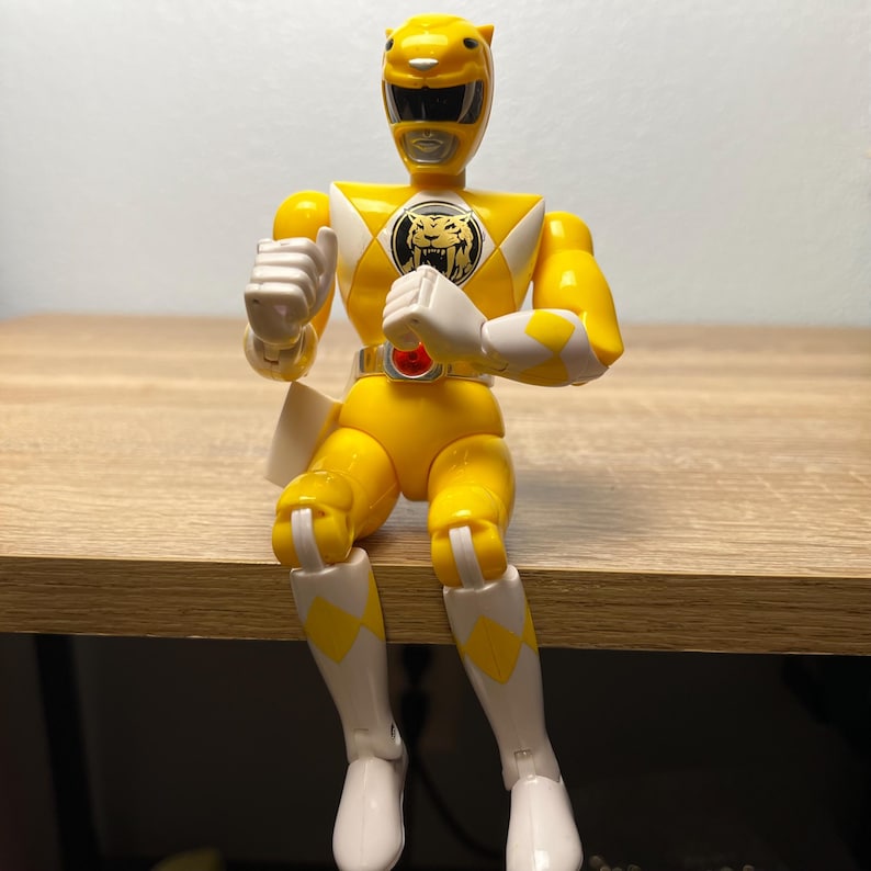 Op de afbeelding: Een gele Power Ranger actiefiguur met witte accenten op de handschoenen en laarzen. De figuur heeft een tijgerembleem op de borst en een gele helm. De figuur zit op een houten oppervlak.