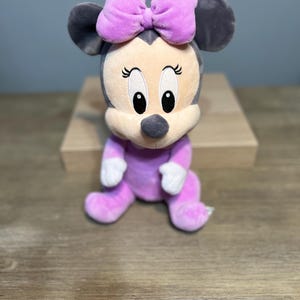 Puede incluir: Mu&ntilde;eca de peluche de Minnie Mouse sentada. La mu&ntilde;eca tiene un atuendo lavanda, un gran lazo lavanda y orejas grises. La cara es beige con ojos negros y una nariz negra. La mu&ntilde;eca est&aacute; sobre una superficie de madera.
