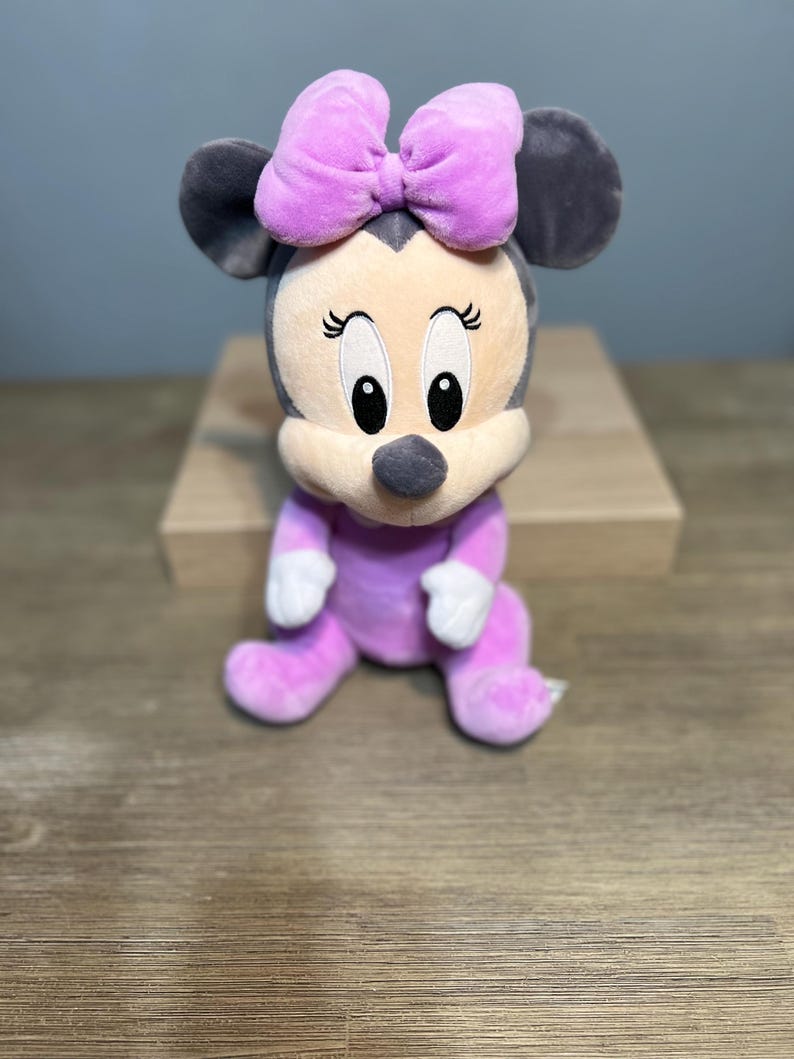 Puede incluir: Mu&ntilde;eca de peluche de Minnie Mouse sentada. La mu&ntilde;eca tiene un atuendo lavanda, un gran lazo lavanda y orejas grises. La cara es beige con ojos negros y una nariz negra. La mu&ntilde;eca est&aacute; sobre una superficie de madera.