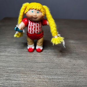 May include: A vintage Cabbage Patch Kids doll with blonde yarn pigtails, blue eyes, and a red and white striped outfit. The doll is wearing red shoes and holding a blue accessory. The doll is a collectible toy.