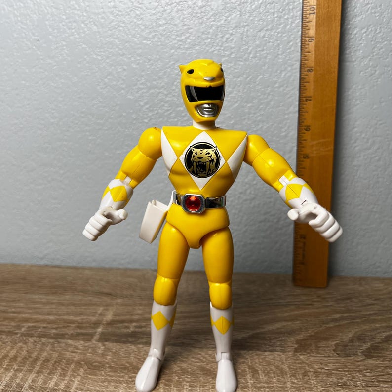 Op de afbeelding: Gele Power Ranger actiefiguur met witte en zwarte accenten. De figuur heeft een tijgerembleem op de borst en een gereedschapsriem. Een houten liniaal staat naast de figuur. De figuur is ongeveer 15 cm hoog.
