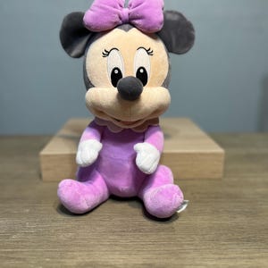 Puede incluir: Mu&ntilde;eca de peluche de Minnie Mouse sentada. La mu&ntilde;eca tiene un atuendo morado claro, un lazo a juego y guantes blancos. La cara de la mu&ntilde;eca es beige con ojos negros y una nariz negra. Las orejas son grises.