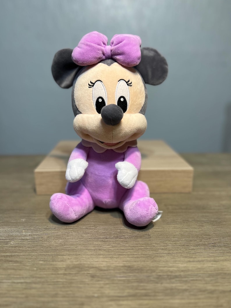 Puede incluir: Mu&ntilde;eca de peluche de Minnie Mouse sentada. La mu&ntilde;eca tiene un atuendo morado claro, un lazo a juego y guantes blancos. La cara de la mu&ntilde;eca es beige con ojos negros y una nariz negra. Las orejas son grises.