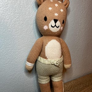 May include: A knitted deer plush toy with brown fur, white spots on the head, and a white muzzle. The deer features black embroidered eyes and a nose, pink cheek accents, and wears light green knit shorts with a drawstring waist detail.