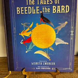 May include: A hardcover book titled "The Tales of Beedle the Bard" by J.K. Rowling. The cover is dark blue with gold lettering and illustrations. The book is illustrated by Lisbeth Zwerger and includes commentary by Albus Dumbledore.
