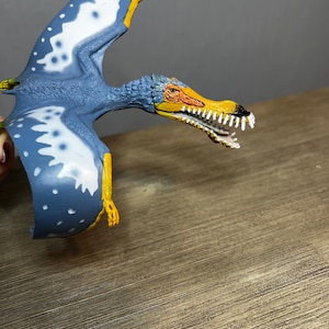 May include: A blue and white toy pterodactyl with yellow accents. The toy has a detailed head with an orange beak and white teeth. The wings are spread, showing white spots. The toy is made of plastic.