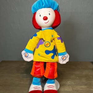 May include: A plush doll of a clown with a blue hat, red hair, and a white face with a red nose. The clown wears a yellow shirt with blue paint splotches, orange shorts, and red shoes. The doll is approximately 14 inches tall.