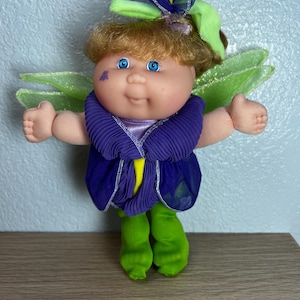 May include: A vintage doll dressed as a fairy, featuring a round face with blue eyes and light brown hair. The doll wears a purple pleated top with a yellow accent, green pants, and green fairy wings. A green bow adorns the doll's head.