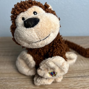 May include: A plush monkey toy featuring dark brown curly fur on its head and back, contrasted with a light beige face, hands, and feet. The monkey has a black nose, a stitched smile, and a small colorful W logo patch on its left foot.
