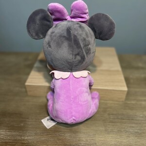 Puede incluir: Un mu&ntilde;eco de peluche de Minnie Mouse, visto desde atr&aacute;s, en posici&oacute;n sentada. El mu&ntilde;eco tiene una cabeza gris con orejas grandes y redondas y un lazo morado. Viste un traje lavanda con un cuello con volantes.