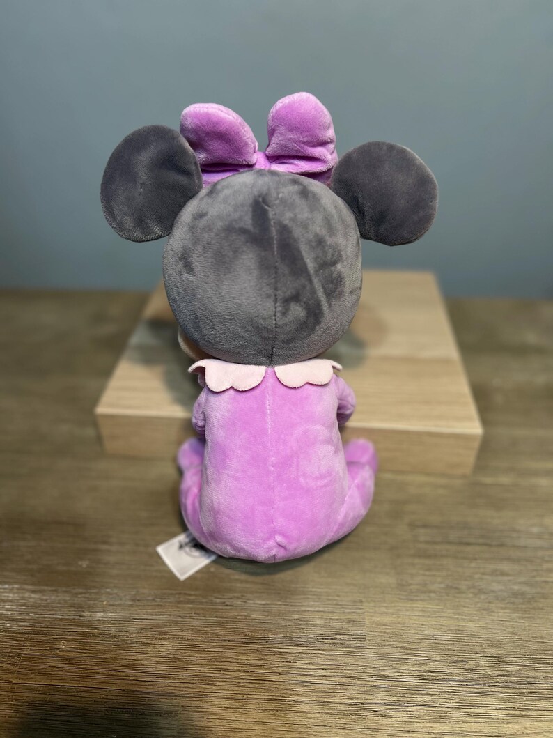 Puede incluir: Un mu&ntilde;eco de peluche de Minnie Mouse, visto desde atr&aacute;s, en posici&oacute;n sentada. El mu&ntilde;eco tiene una cabeza gris con orejas grandes y redondas y un lazo morado. Viste un traje lavanda con un cuello con volantes.