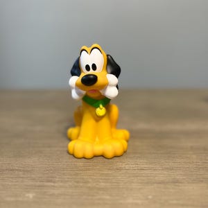 Puede incluir: Una figura amarilla de Pluto con orejas negras y una nariz negra. El perro lleva un collar verde con una etiqueta dorada y sostiene un hueso blanco en la boca. La figura está sobre una superficie de madera.