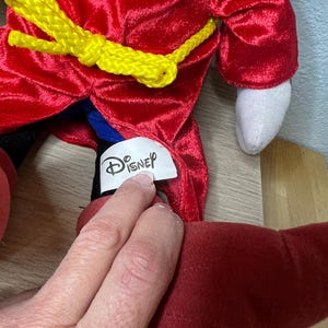 K&ouml;nnte beinhalten: Ein Pl&uuml;schtier mit einem roten, samtartigen Mantel und einem gelben Seilg&uuml;rtel. Das Spielzeug hat eine wei&szlig;e Hand und einen roten Fu&szlig;. Das Disney-Logo ist auf einem Etikett zu sehen.