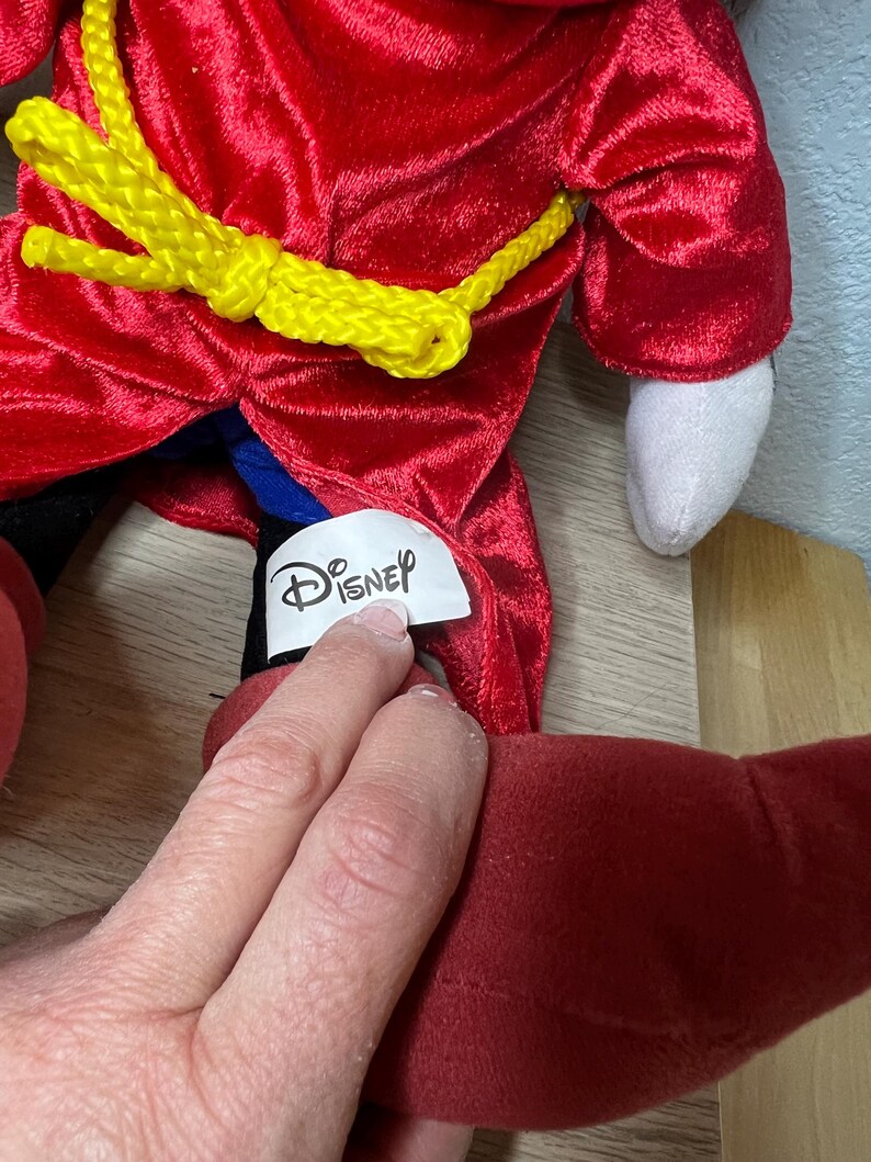 K&ouml;nnte beinhalten: Ein Pl&uuml;schtier mit einem roten, samtartigen Mantel und einem gelben Seilg&uuml;rtel. Das Spielzeug hat eine wei&szlig;e Hand und einen roten Fu&szlig;. Das Disney-Logo ist auf einem Etikett zu sehen.