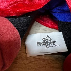 K&ouml;nnte beinhalten: Nahaufnahme eines Pl&uuml;schtiers mit rotem, schwarzem und blauem Stoff. Ein wei&szlig;es Etikett mit dem Text "Toy Factory" und "San Antonio, TX" ist sichtbar.