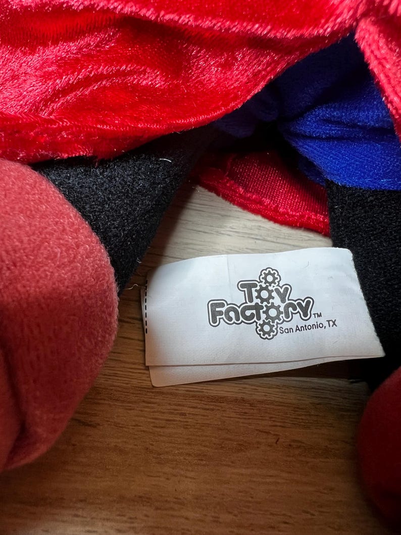 K&ouml;nnte beinhalten: Nahaufnahme eines Pl&uuml;schtiers mit rotem, schwarzem und blauem Stoff. Ein wei&szlig;es Etikett mit dem Text "Toy Factory" und "San Antonio, TX" ist sichtbar.