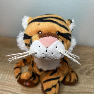 May include: A plush tiger toy featuring orange fur with black stripes, a white muzzle, a pink nose, and long white whiskers. The stuffed animal has small black eyes and white tufts of fur on the sides of its head, with a small colorful circular emblem on one paw.